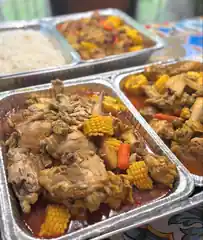 Large catering trays filled with stewed chicken, corn, vegetables, and sauce ready for festival service.